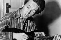 A black and white photograph of American folk musician Woody Guthrie sitting indoors, looking upward and to his left. He is wearing a plaid button-down shirt and a dark, flat cap on his curly hair. He is holding and playing an acoustic guitar. On the top upper bout of the guitar's body, a prominent rectangular sticker reads in bold, hand-lettered capital letters: "THIS MACHINE KILLS FASCISTS". The background features a simple paneled wooden door with a round doorknob on the left and a plain wall casting a shadow on the right.